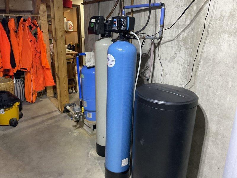 Conducted comprehensive inspection of residential water treatment system, including multiple filtration tanks (two blue cylindrical, one gray/beige, and one black brine tank) with digital control panels and associated PVC plumbing. Examined drainage components, including main white PVC drain pipe with elbow joint and connected metallic piping, documenting current condition with photos for reference.